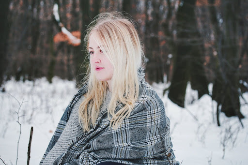 A blonde woman sitting outside in the snow, wrapped in a blanket.