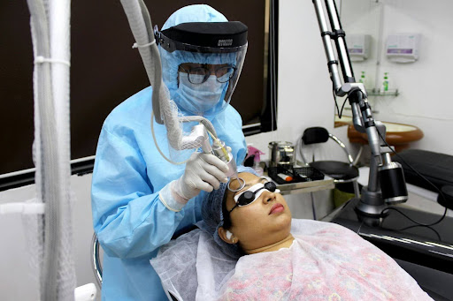 A woman with black goggles on getting hyperpigmentation laser treatment. THe cosmetic surgeon is wearing a blue bodysuit and a plastic face shield.